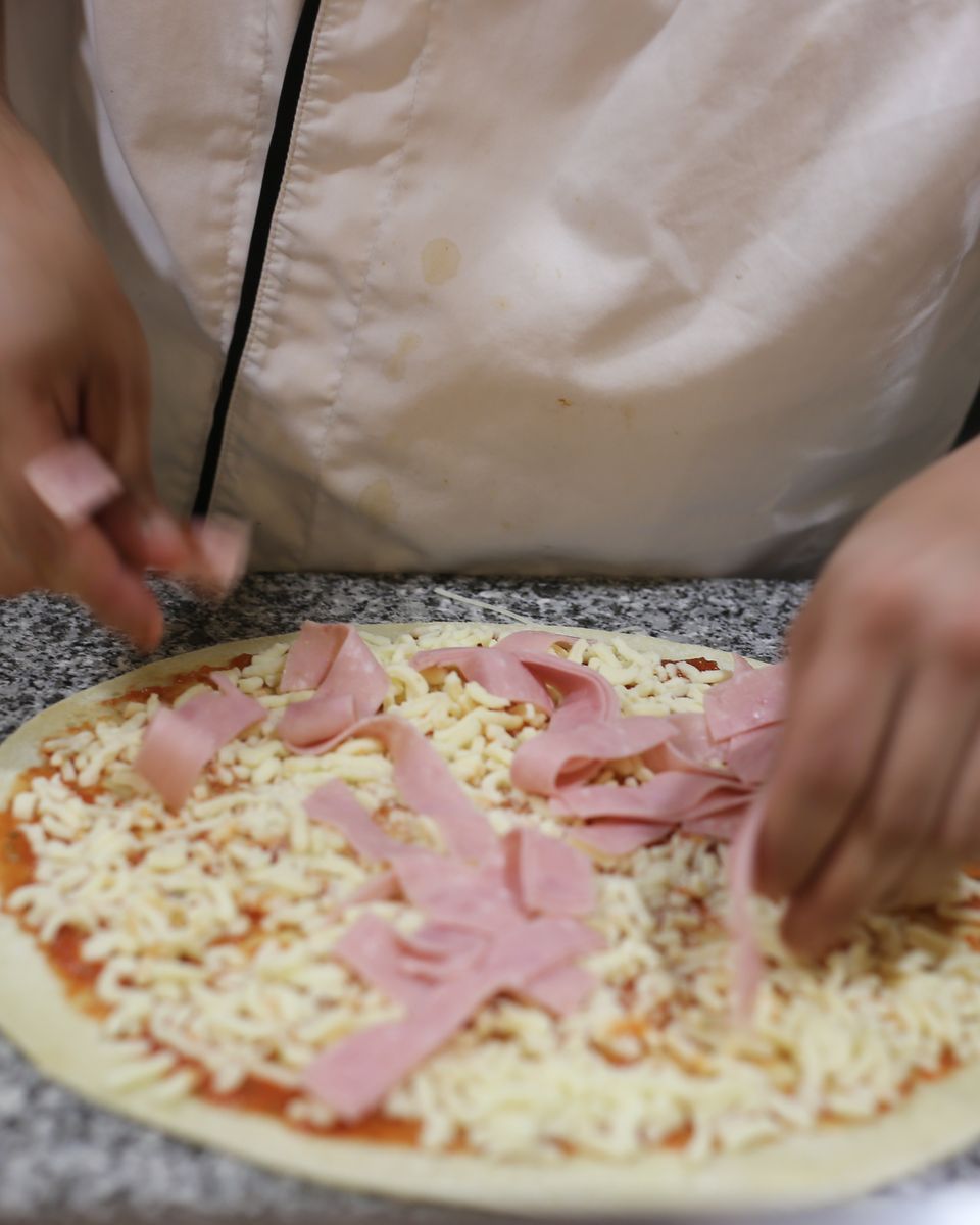 Pizza a ser coberta de queijo e fiambre na bancada